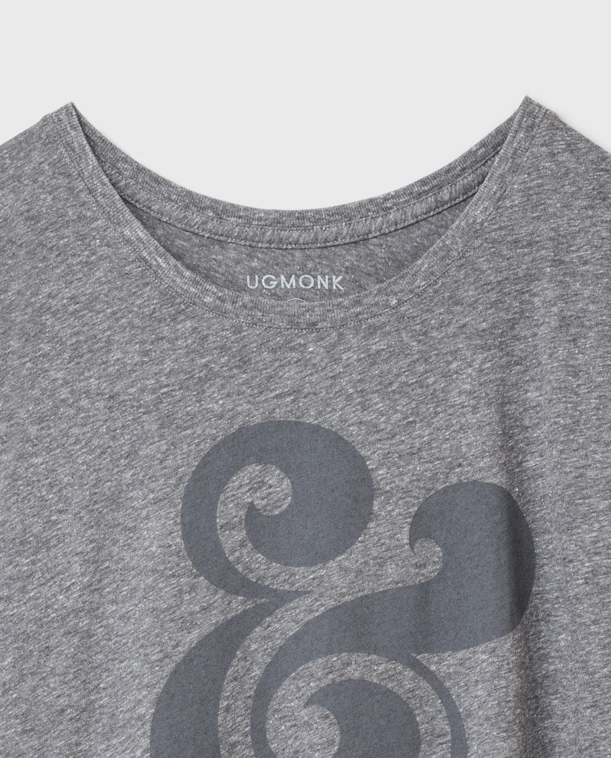 A heather gray t-shirt with UGMONK printed inside the collar and a large, stylized dark gray ampersand graphic on the front. The shirt has a simple round neckline.
