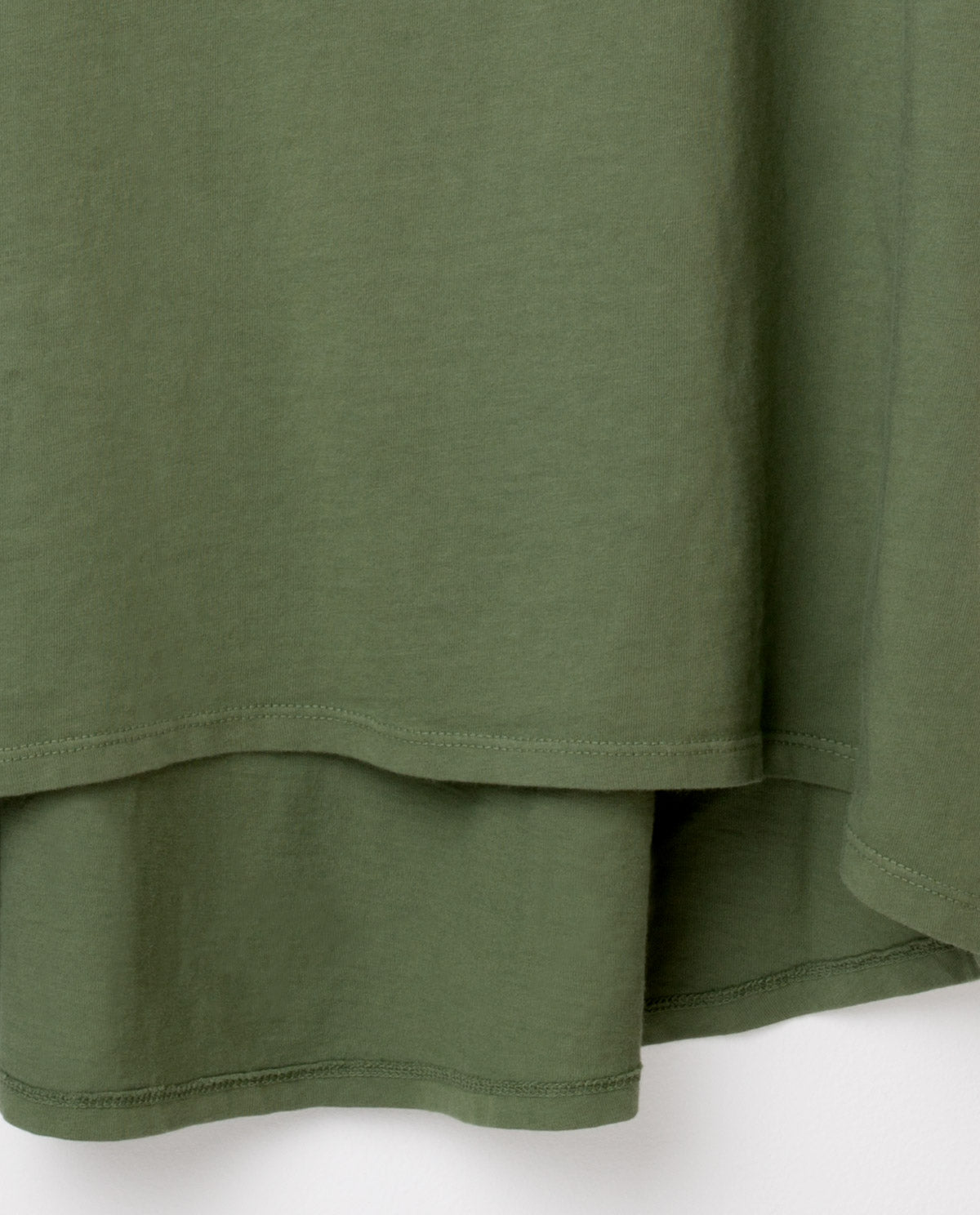 Close-up of a green fabric with a layered, asymmetrical hem, likely part of a garment such as a dress or skirt. The fabric has a smooth texture and the stitching is visible along the edge.