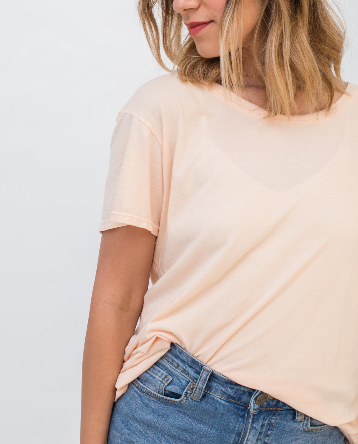 A woman with shoulder-length blonde hair wears a light peach t-shirt and blue jeans, standing against a plain white background. Her face is partially visible and she is slightly turned to the side.