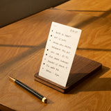 A handwritten to-do list sits on the sunlit wooden table in a walnut card holder beside a refillable pen—tasks like writing a draft and attending a 1 pm meeting are neatly organized in The Analog Bundle (Walnut).