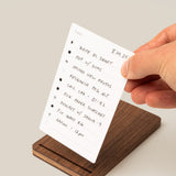 A hand holds a white notecard listing tasks above The Analog Bundle (Walnut), a sleek card holder. The to-do list includes writing, uploading photos, research, calls, and meetings. The scene has a clean, light background.