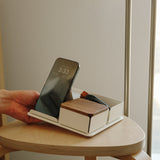 A hand places a smartphone onto the Gather 2x2 Organizer Set (White) tray on a wooden stool. The tray also holds a closed wooden box and small items, positioned near a window with natural light.