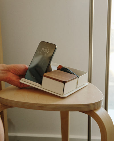 A hand places a smartphone onto the Gather 2x2 Organizer Set (White) tray on a wooden stool. The tray also holds a closed wooden box and small items, positioned near a window with natural light.