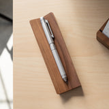 The Craft Design Technology Multifunctional Pen in silver sits on a rectangular wooden holder atop a light wood desk, with soft sunlight casting gentle shadows across the surface.