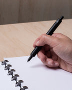 A hand holds the HMM Ballpoint Pen (Black) above a dotted notebook with silver disc bindings, on a light wooden desk.
