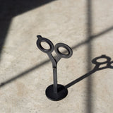 A pair of HMM Scissors (Black) stands upright on a magnetic metal base, casting a distinct shadow on a sunlit surface with intersecting lines of light and shadow.