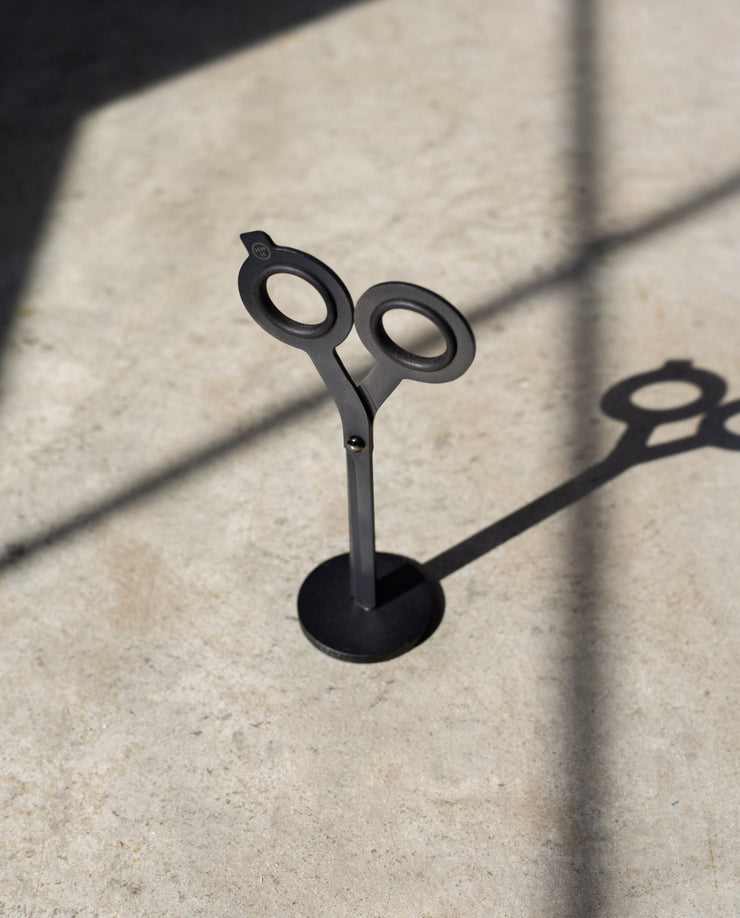 A pair of HMM Scissors (Black) stands upright on a magnetic metal base, casting a distinct shadow on a sunlit surface with intersecting lines of light and shadow.