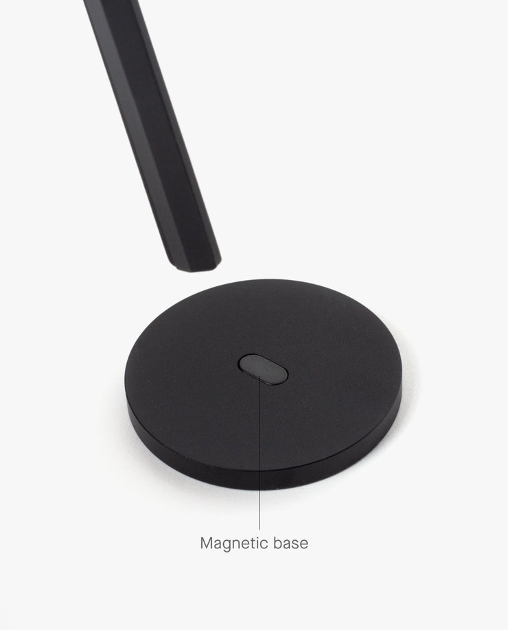 A round, black magnetic metal base is displayed on a white background, with HMM Scissors (Black) hovering above it. A label points to the base, reading "Magnetic base.