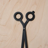 HMM Scissors (Black) with round handles and a minimalist design rest closed on a light wood surface, handles up. A magnetic metal base complements their sleek look.