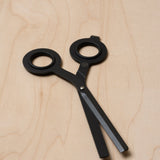 The HMM Scissors (Black) feature round handles and two blunt, flat blades. Decorative and non-functional, they rest on a light wood surface and can be displayed on a magnetic metal base.