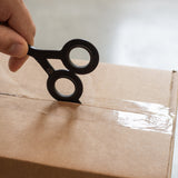 A hand uses HMM Scissors (Black), a small circular cutting tool with a built-in box cutter, to slice through clear tape sealing a brown cardboard box.