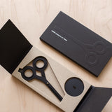 A pair of HMM Scissors (Black) with round handles is shown in an open black box featuring minimalist packaging and a magnetic base, while a similar closed box rests nearby on a light wood surface.