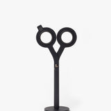 The HMM Scissors (Black) is a modern, minimalist desk sculpture with circular handles and a sleek geometric design, standing upright on a magnetic metal base.