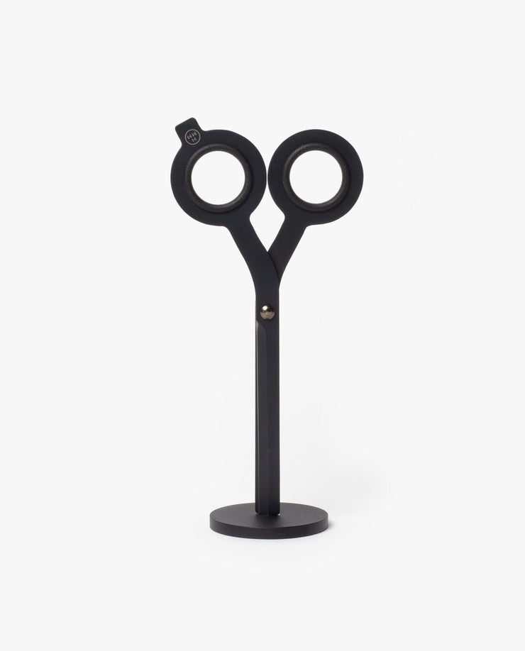 The HMM Scissors (Black) is a modern, minimalist desk sculpture with circular handles and a sleek geometric design, standing upright on a magnetic metal base.