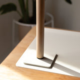 A close-up of the Gather Headphone Stand (1x1 - White/Maple), featuring solid maple wood on a white powder-coated steel base, sits on the corner of a light wood desk with sunlight and a blurred potted plant in the background.