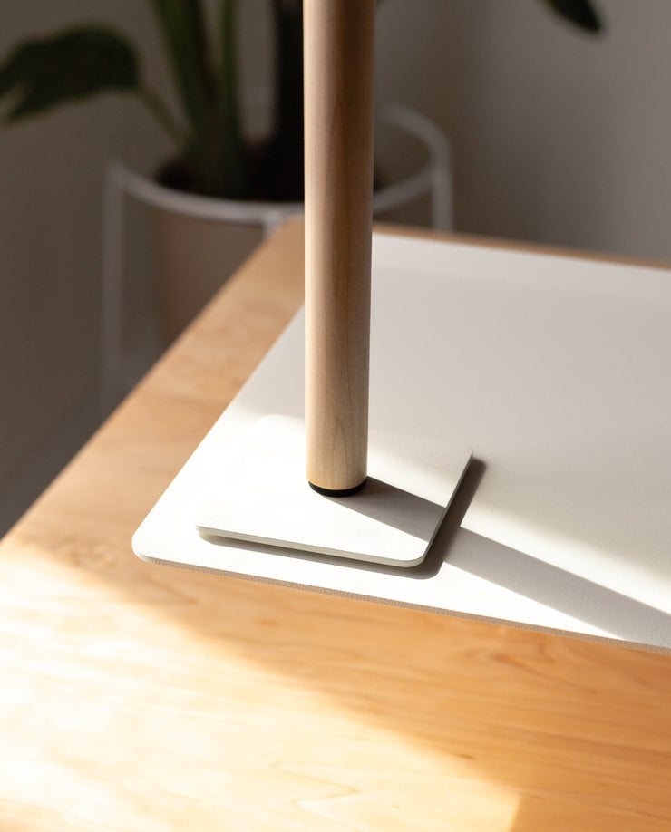 A close-up of the Gather Headphone Stand (1x1 - White/Maple), featuring solid maple wood on a white powder-coated steel base, sits on the corner of a light wood desk with sunlight and a blurred potted plant in the background.