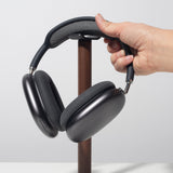 A hand places over-ear black headphones onto the Gather Headphone Stand (1x1 - Black/Walnut), which features a solid walnut wood post and a powder-coated steel base, set on a white surface against a plain light background.