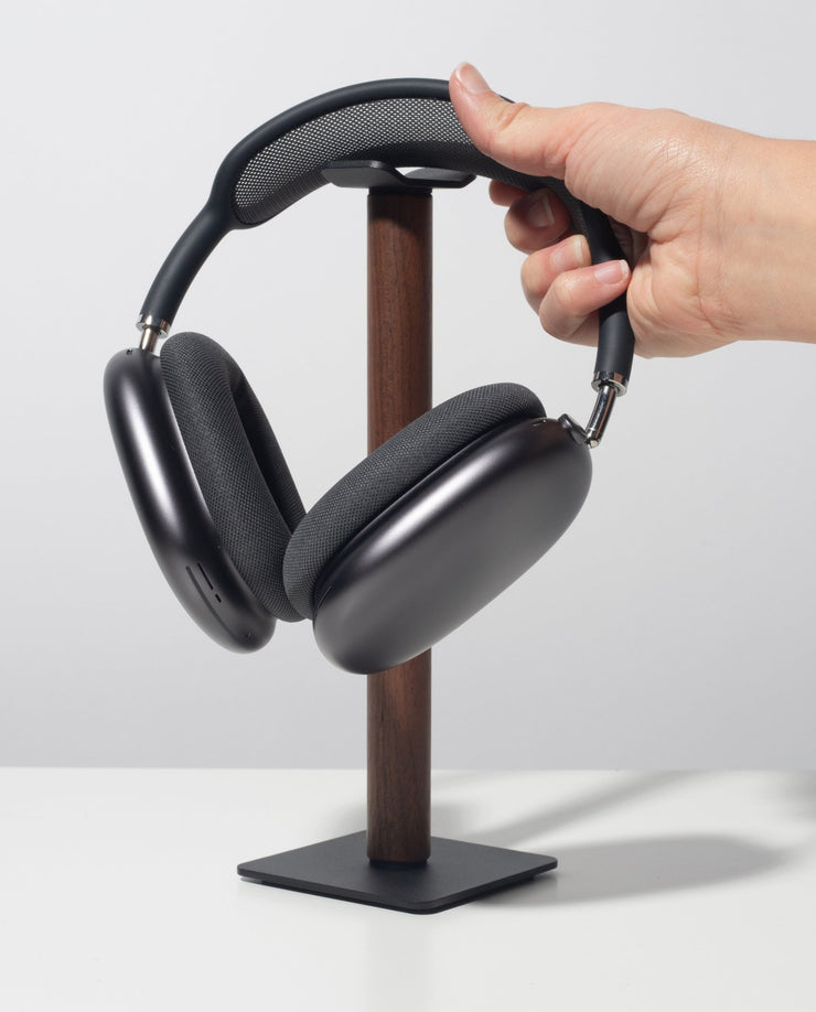 A hand places over-ear black headphones onto the Gather Headphone Stand (1x1 - Black/Walnut), which features a solid walnut wood post and a powder-coated steel base, set on a white surface against a plain light background.