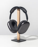 The Gather Headphone Stand (1x1 - Black/Maple), featuring a solid maple support and a powder coated steel square base, displays a pair of black over-ear headphones on a white surface against a plain light gray background.