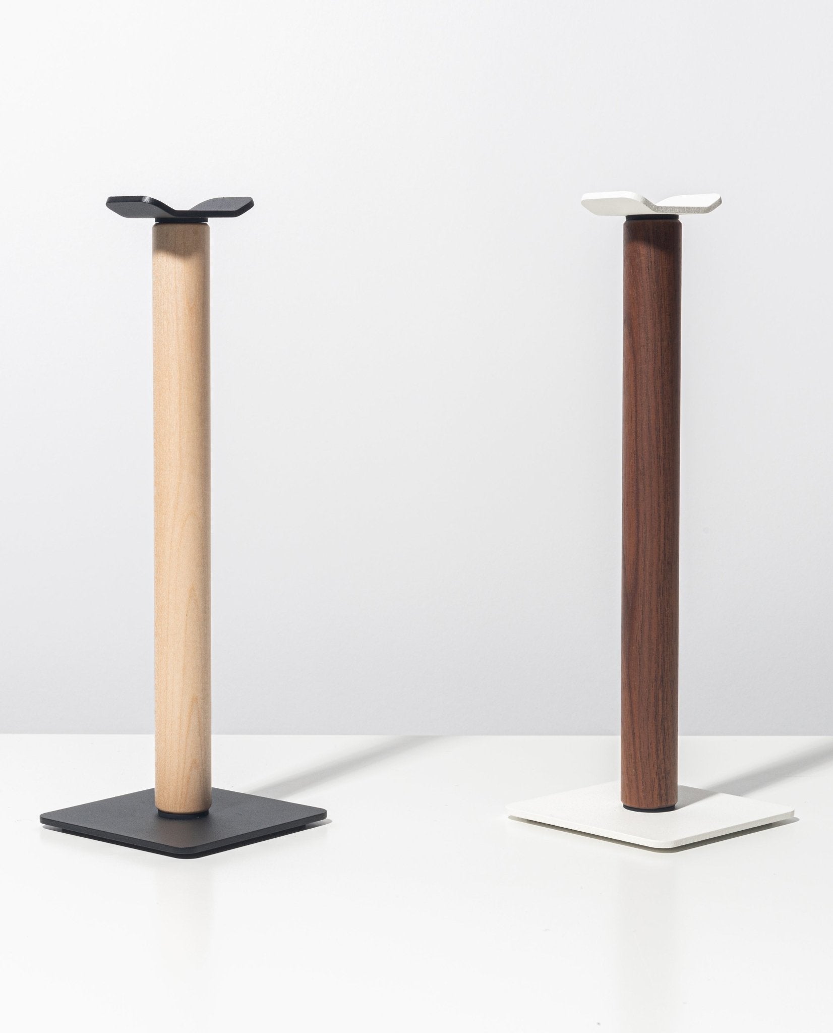 Two modern, minimalist Gather Headphone Stands (1x1 - Black/Maple) stand side by side on a white surface. One features a solid maple wood base with black powder coated steel accents, while the other pairs a dark wood base with sleek white details.