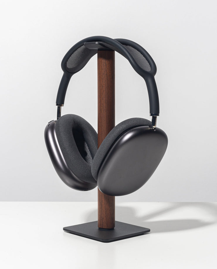Sleek, over-ear black headphones with a mesh headband rest on the Gather Headphone Stand (1x1 - Black/Walnut), crafted from solid wood and powder-coated steel, displayed against a plain white background.