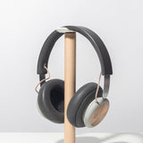 The Gather Headphone Stand (1x1 - White/Maple) holds over-ear headphones with gray ear cups and copper accents. The stand has a solid maple support and a square white powder-coated steel base, displayed on a plain white background.