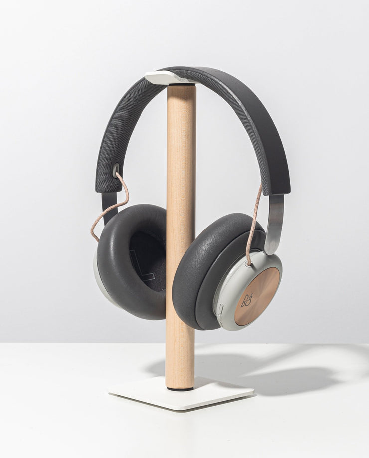 The Gather Headphone Stand (1x1 - White/Maple) holds over-ear headphones with gray ear cups and copper accents. The stand has a solid maple support and a square white powder-coated steel base, displayed on a plain white background.