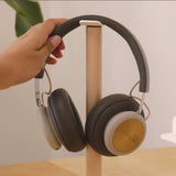 A hand places a pair of over-ear headphones with gold accents onto the Gather Headphone Stand (1x1 - White/Maple), which features solid wood and powder coated steel, set on a light-colored desk.
