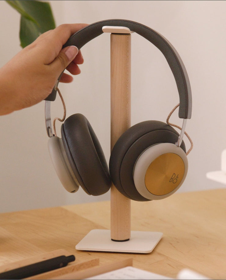 A hand places a pair of over-ear headphones with gold accents onto the Gather Headphone Stand (1x1 - White/Maple), which features solid wood and powder coated steel, set on a light-colored desk.