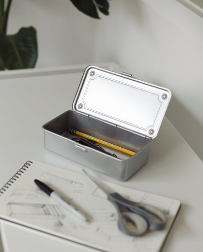 A Toyo Steel Stackable Storage Box T-190 (Silver) with its lid open sits on a white desk, holding a pencil and pen. In front are an open sketchbook, black marker, and scissors. A plant is in the background. Made in Japan.