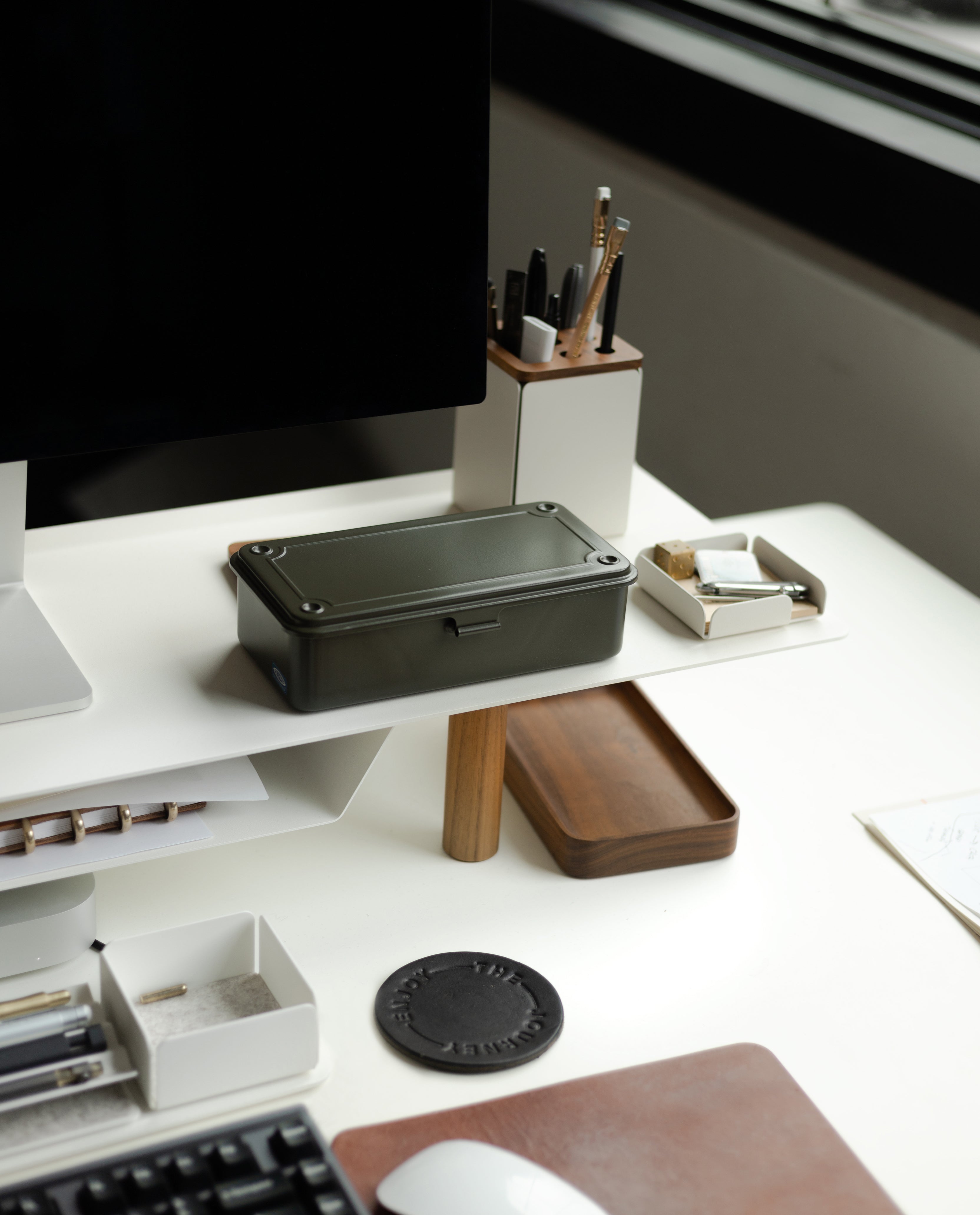 A modern desk features a computer, pen holder, trays with office supplies, and a Toyo Steel Stackable Storage Box T-190 (Military Green) made in Japan, all arranged near a large window that provides natural light.