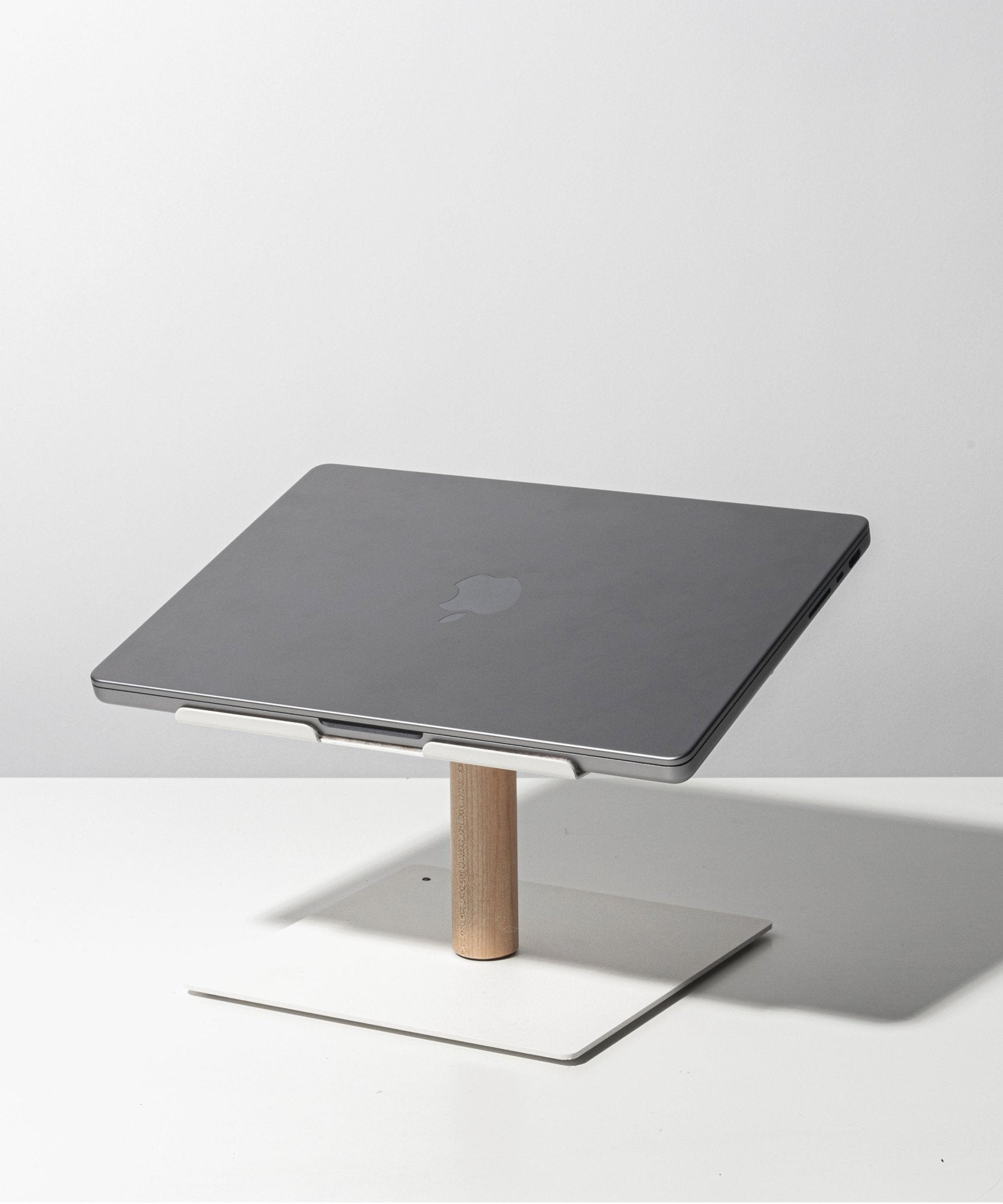 A closed gray laptop sits on the Gather Laptop Stand (White/Maple), featuring a minimalist white and maple wood design, placed on a white surface against a light gray background.
