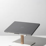 A closed gray laptop sits on the Gather Laptop Stand (White/Maple), featuring a minimalist white and maple wood design, placed on a white surface against a light gray background.