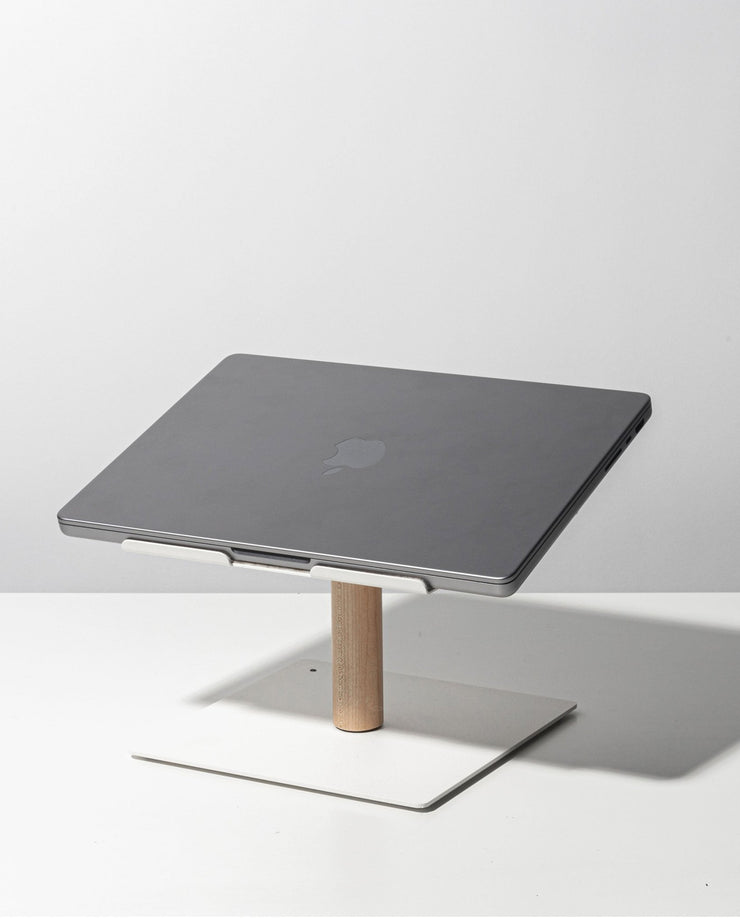 A closed gray laptop sits on the Gather Laptop Stand (White/Maple), featuring a minimalist white and maple wood design, placed on a white surface against a light gray background.