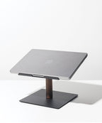 A closed silver Apple laptop sits on the Gather Laptop Stand (Black/Walnut), which has a black base and walnut support, placed on a white surface against a plain white background.