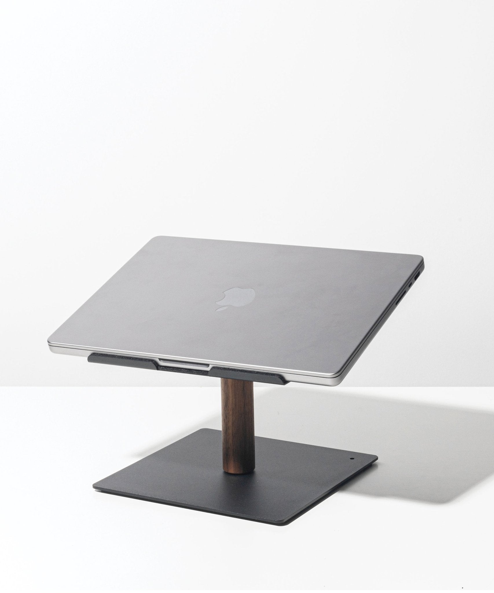 A closed silver Apple laptop sits on the Gather Laptop Stand (Black/Walnut), which has a black base and walnut support, placed on a white surface against a plain white background.