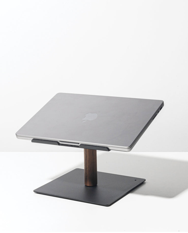 A closed silver Apple laptop sits on the Gather Laptop Stand (Black/Walnut), which has a black base and walnut support, placed on a white surface against a plain white background.
