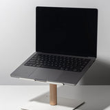 A black-screen laptop rests on the Gather Laptop Stand (White/Maple), featuring a maple rod and white base, placed on a white table against a plain light background.