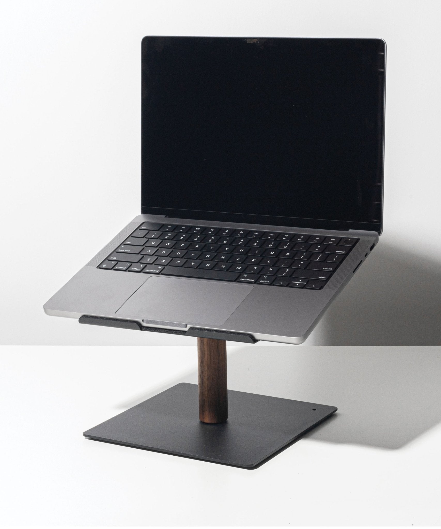 A Gather Laptop Stand (Black/Walnut) holds an open laptop with a black keyboard; the stand features a dark walnut stem and flat metal base, set on a white surface against a plain background.