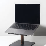 A Gather Laptop Stand (Black/Walnut) holds an open laptop with a black keyboard; the stand features a dark walnut stem and flat metal base, set on a white surface against a plain background.