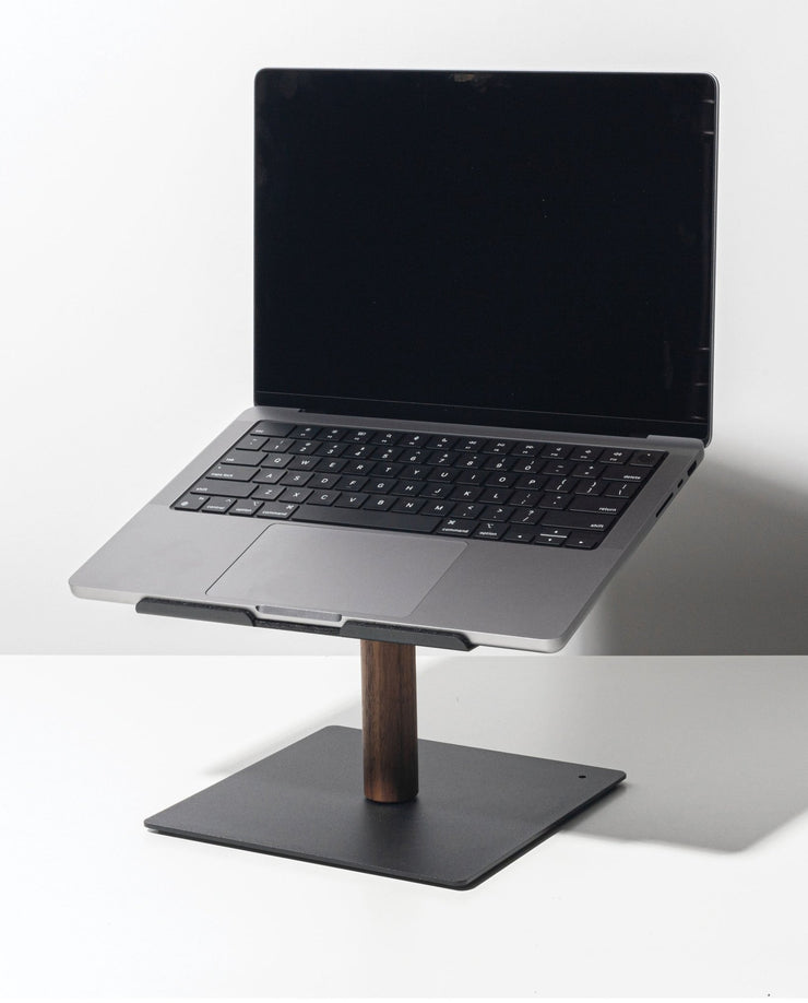 A Gather Laptop Stand (Black/Walnut) holds an open laptop with a black keyboard; the stand features a dark walnut stem and flat metal base, set on a white surface against a plain background.
