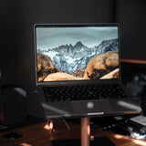 A laptop displaying a mountain landscape is placed on the Gather Laptop Stand (Black/Walnut) on a desk, sunlight streaming across the surface.