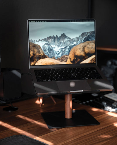 A laptop displaying a mountain landscape is placed on the Gather Laptop Stand (Black/Walnut) on a desk, sunlight streaming across the surface.