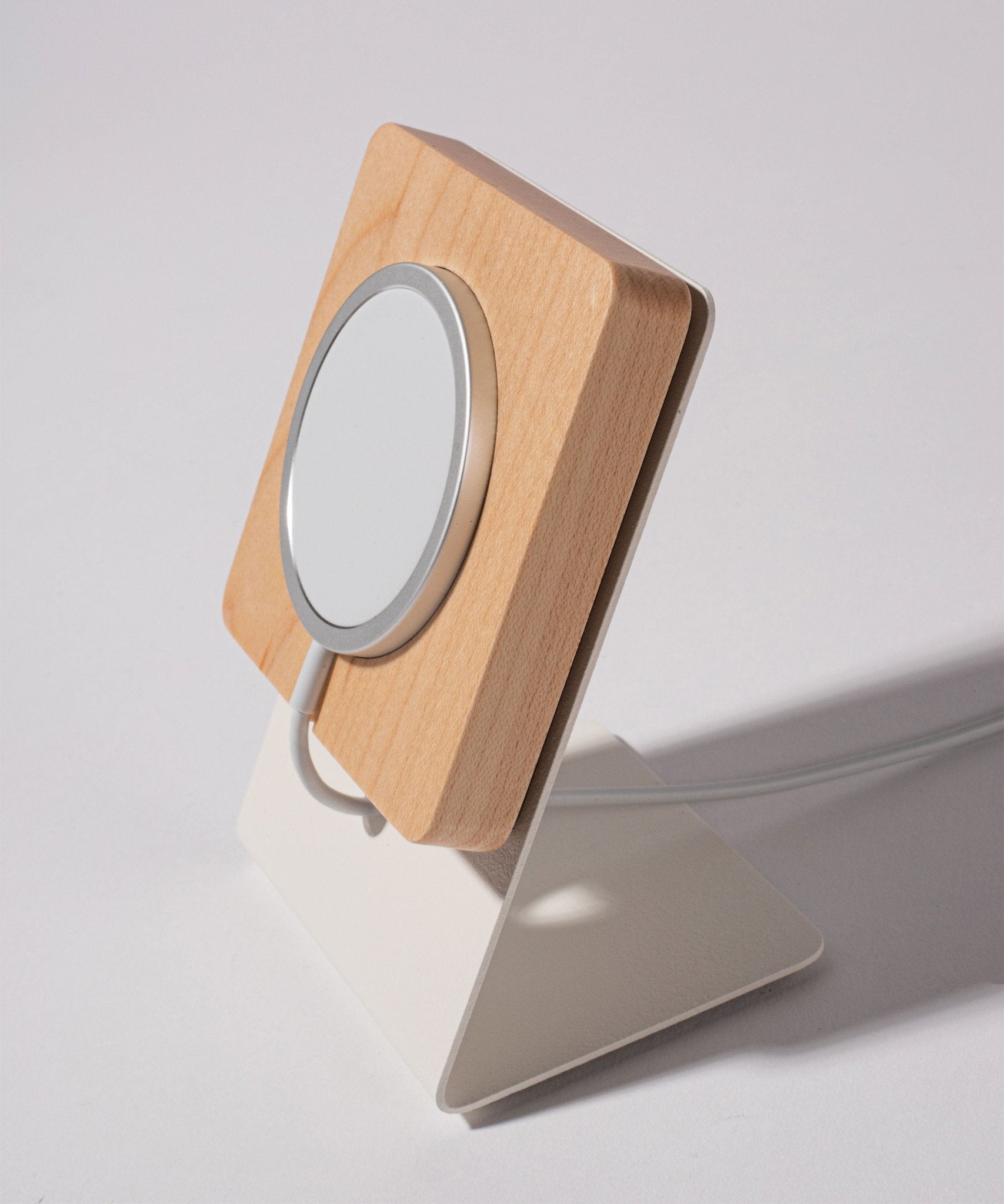 The Gather MagSafe Phone Stand (1x1 - White/Maple), featuring a combination of wood and metal with a circular MagSafe pad, functions as a stylish wireless charging stand. It rests on a white angled base against a plain light background, with the charging cable subtly connected at the back.