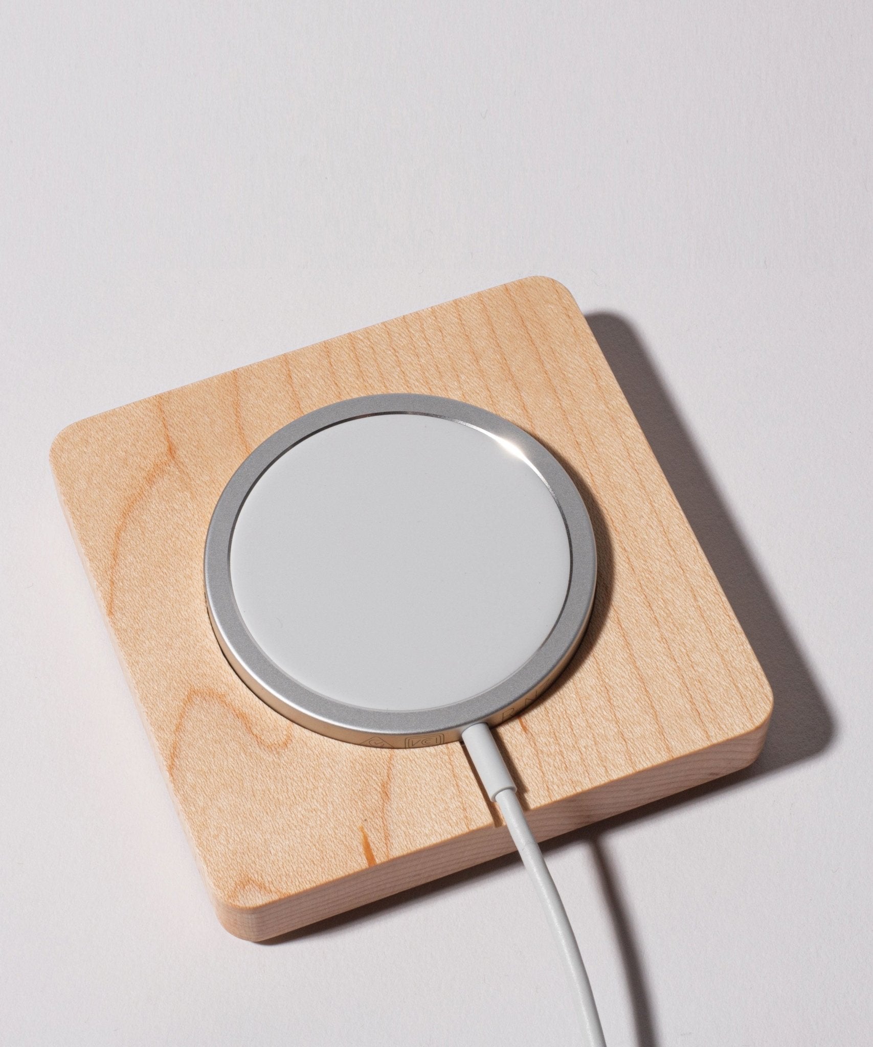 The Gather MagSafe Phone Stand (1x1 - White/Maple), featuring a round wireless charging pad with a white surface and silver edge, rests on a square maple wood base. A white cable extends from the charging pad, all set against a plain white background.