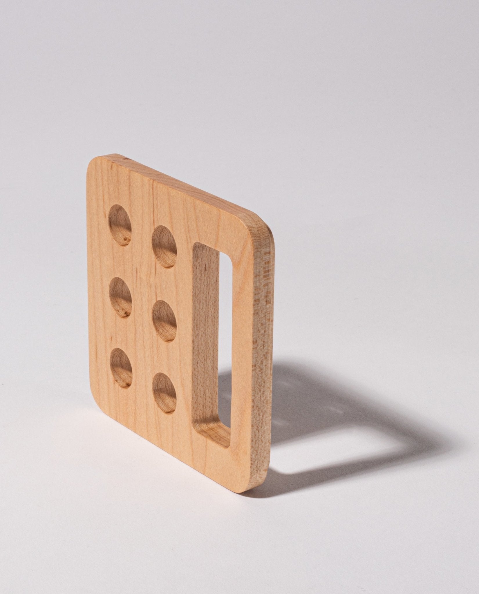 A square maple Gather Pencil Cup Lid, handcrafted in Pennsylvania from solid wood, features five round holes on one side and a large rectangular cutout, casting a shadow on a light surface.