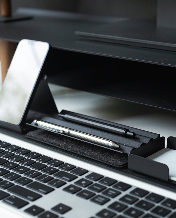 A smartphone, pens, and a stylus are neatly arranged in a black Gather Zigzag Tray (2x1) made from powder-coated steel beside a computer keyboard on a modern, minimal workspace.