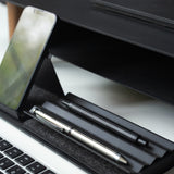 Close-up of a workspace featuring a smartphone on a stand, two pens in the Gather Zigzag Tray (2x1 - Black), and part of a laptop keyboard on a white desk.