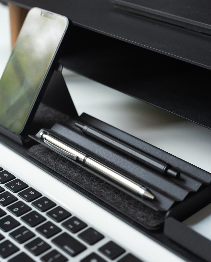 Close-up of a workspace featuring a smartphone on a stand, two pens in the Gather Zigzag Tray (2x1 - Black), and part of a laptop keyboard on a white desk.