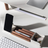 A minimalist white desk setup includes a wireless keyboard, silver pen on a walnut pen holder, smartphone, and the Gather Wood Zigzag Tray (2x1 - Walnut) holding keys and USB drives—all neatly arranged under bright lighting.